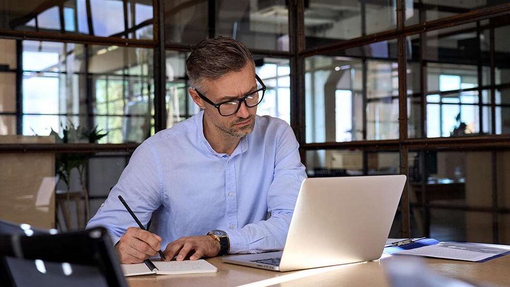 Man sitting at a table working on a computer and taking notes